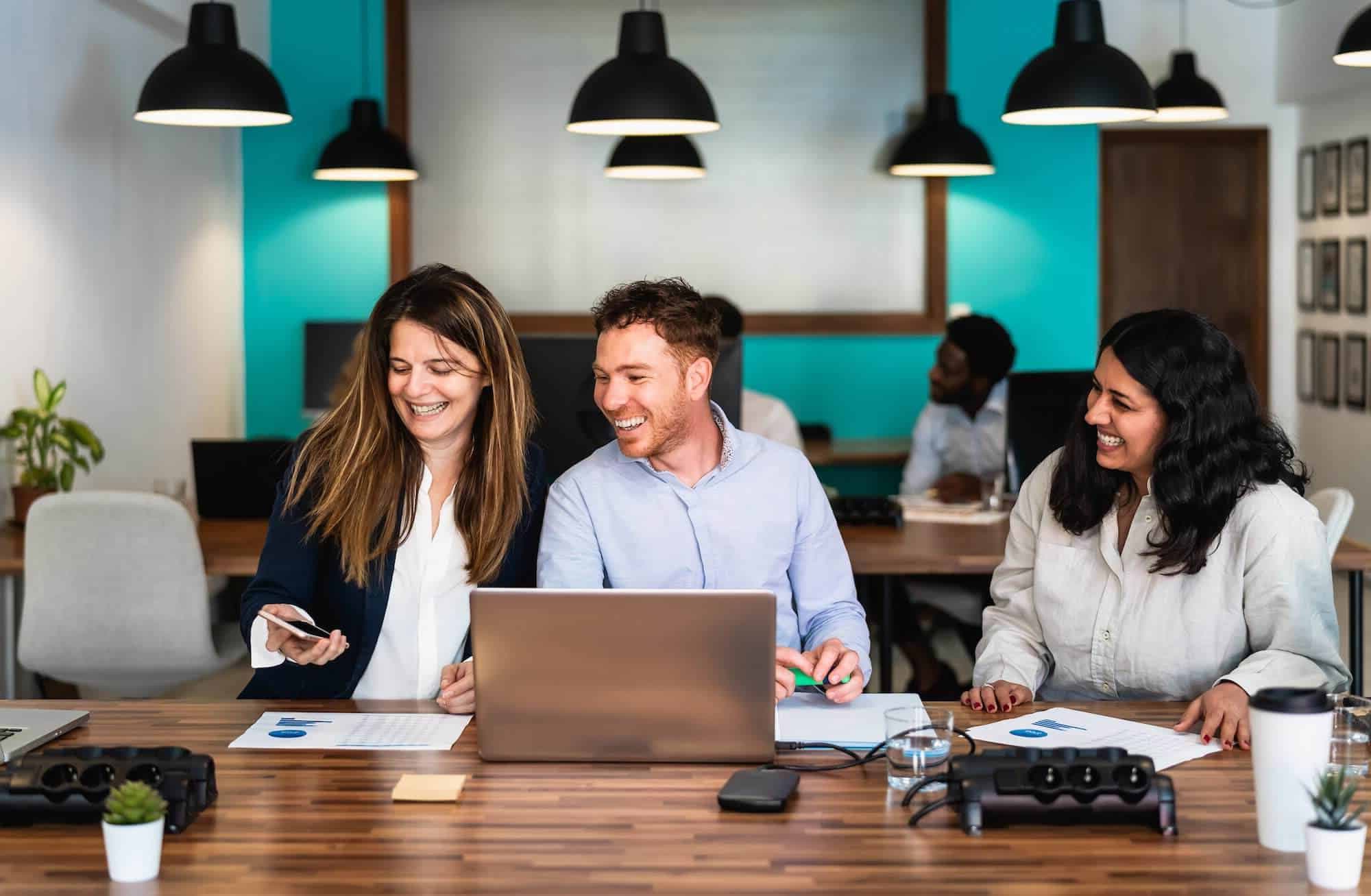 Team collaborating in a modern co-working space, smiling and engaged in discussion with laptops, documents, and a creative atmosphere.